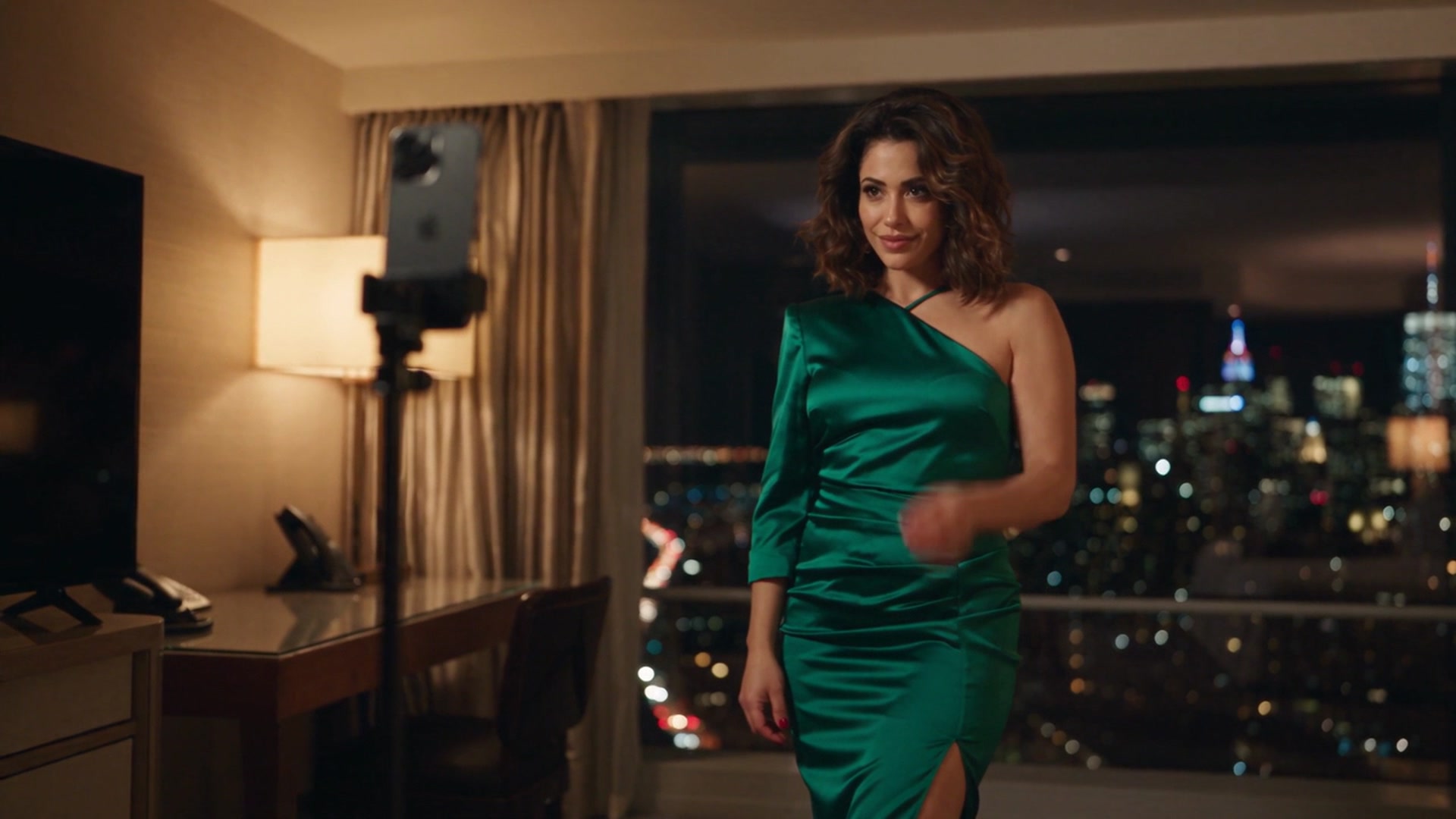 Amal wearing a green satin one-shoulder dress with a thigh-high slit, walking through a hotel room at night with a smartphone recording on a tripod and a city skyline visible through the window behind her.