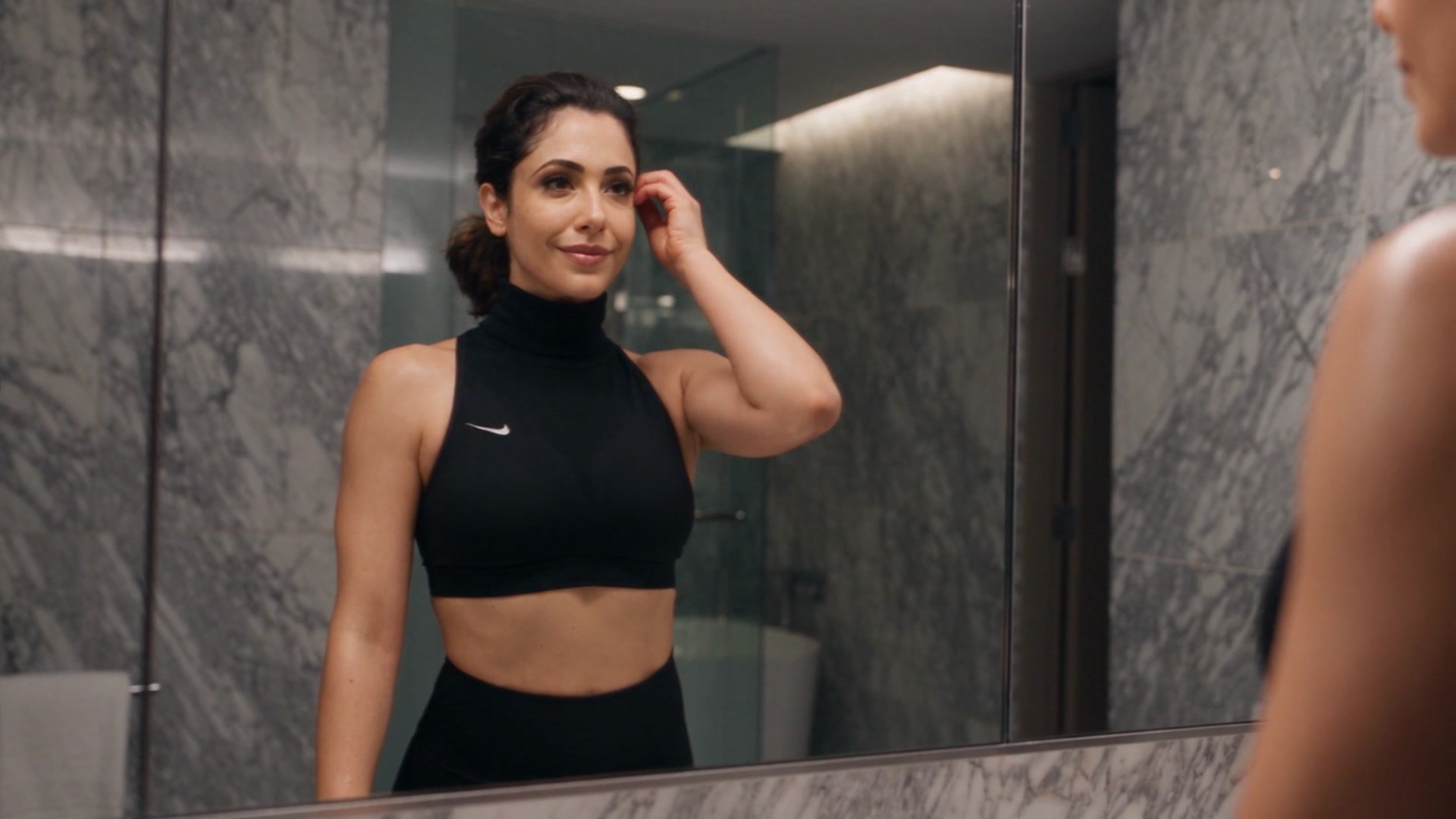 Amal wearing a black high-neck sleeveless athletic top and leggings, standing in a marble-tiled bathroom and tucking her hair back while looking at her reflection in the mirror.