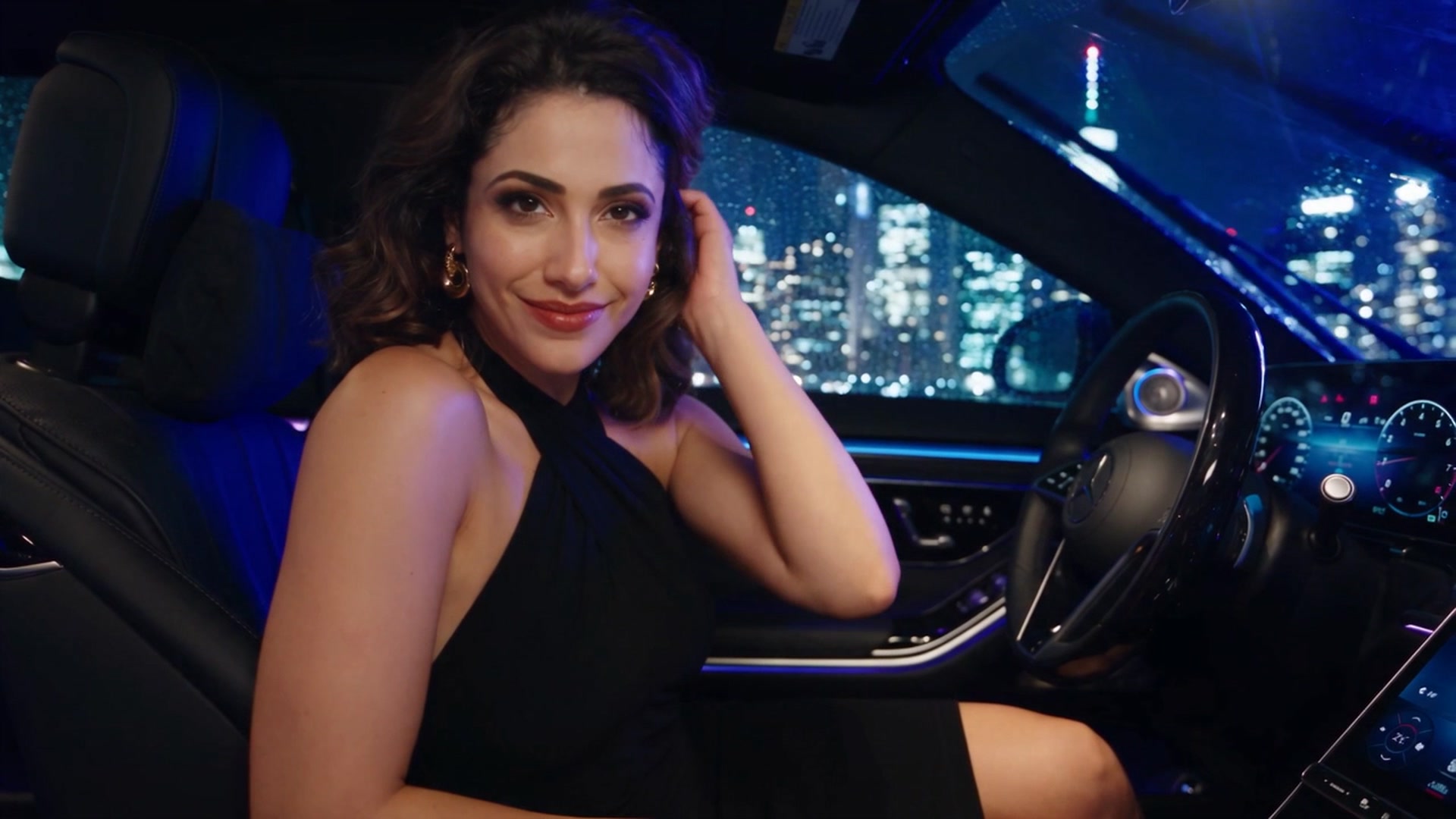 Amal wearing a black halter dress and gold earrings, seated in a car at night with one hand raised to her hair and a lit-up city skyline visible through the window.