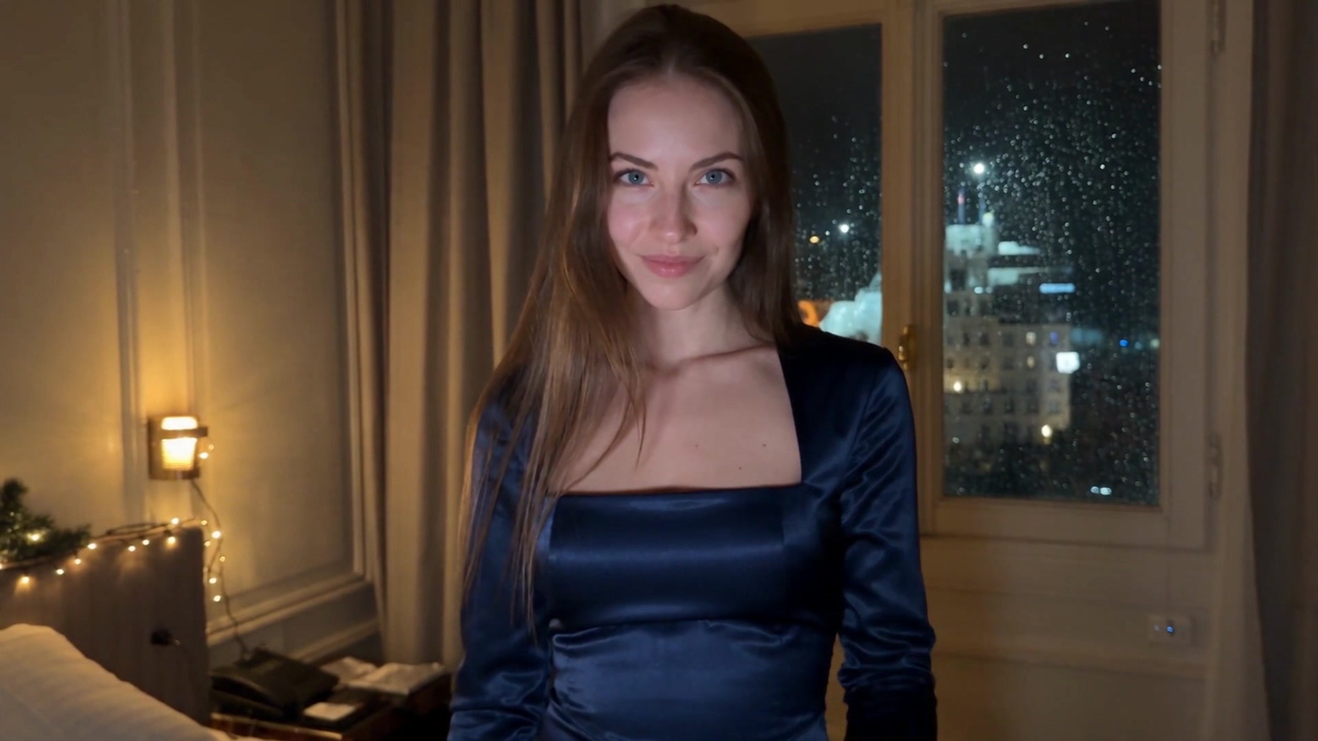 Amina wearing a navy satin off-shoulder-style dress with a square neckline, standing in a softly lit bedroom with sheer curtains and a rain-streaked window overlooking a city skyline at night.