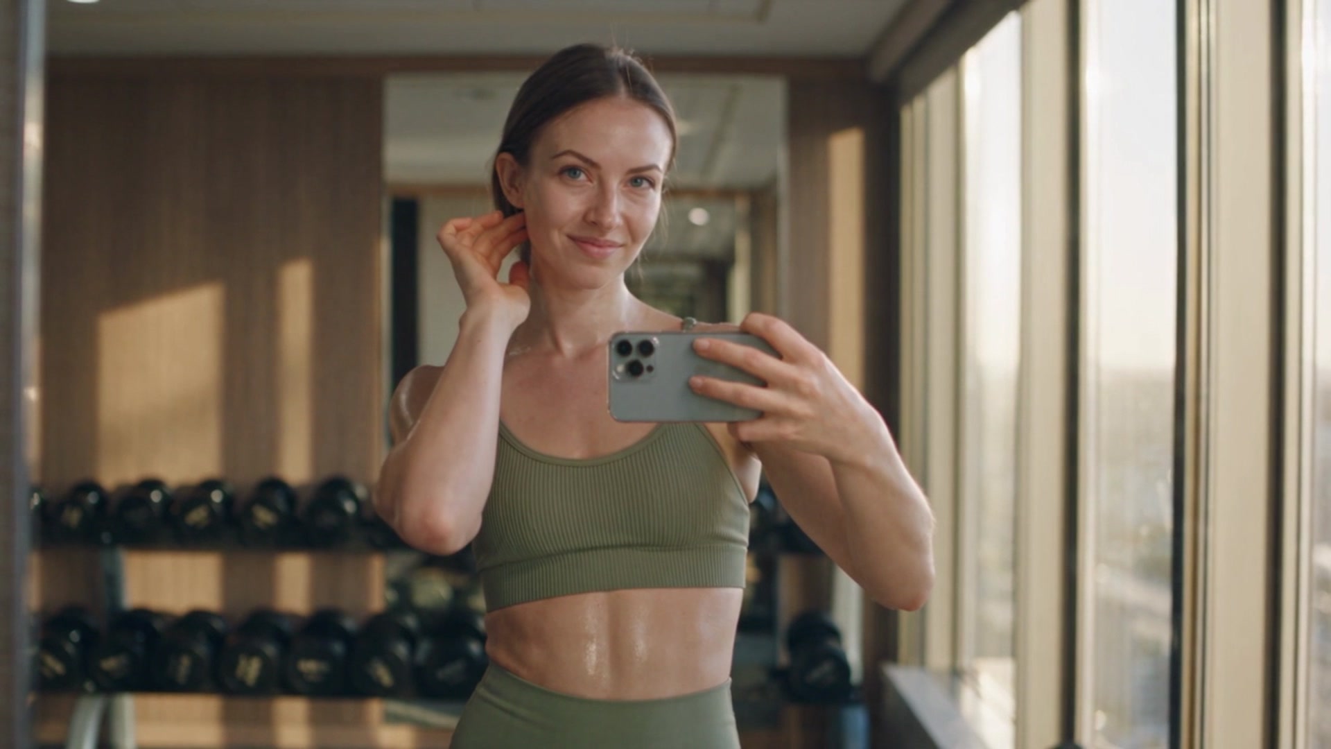 Amina wearing an olive green ribbed sports bra and matching leggings, taking a mirror selfie with a smartphone in a home gym lined with dumbbell racks and floor-to-ceiling windows.