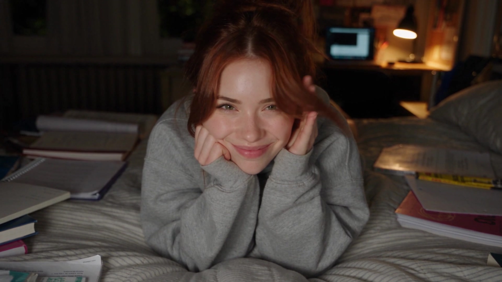 Elena wearing a heather gray oversized sweatshirt, lying on her stomach on a bed surrounded by notebooks and textbooks, resting her chin on her hands and smiling at the camera in a cozy, lamp-lit room.