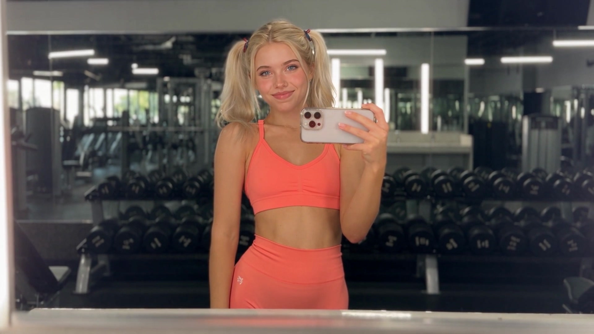 Emely in a coral sports bra and matching high-waisted leggings with hair in twin ponytails, taking a mirror selfie with a phone in a gym surrounded by dumbbell racks.