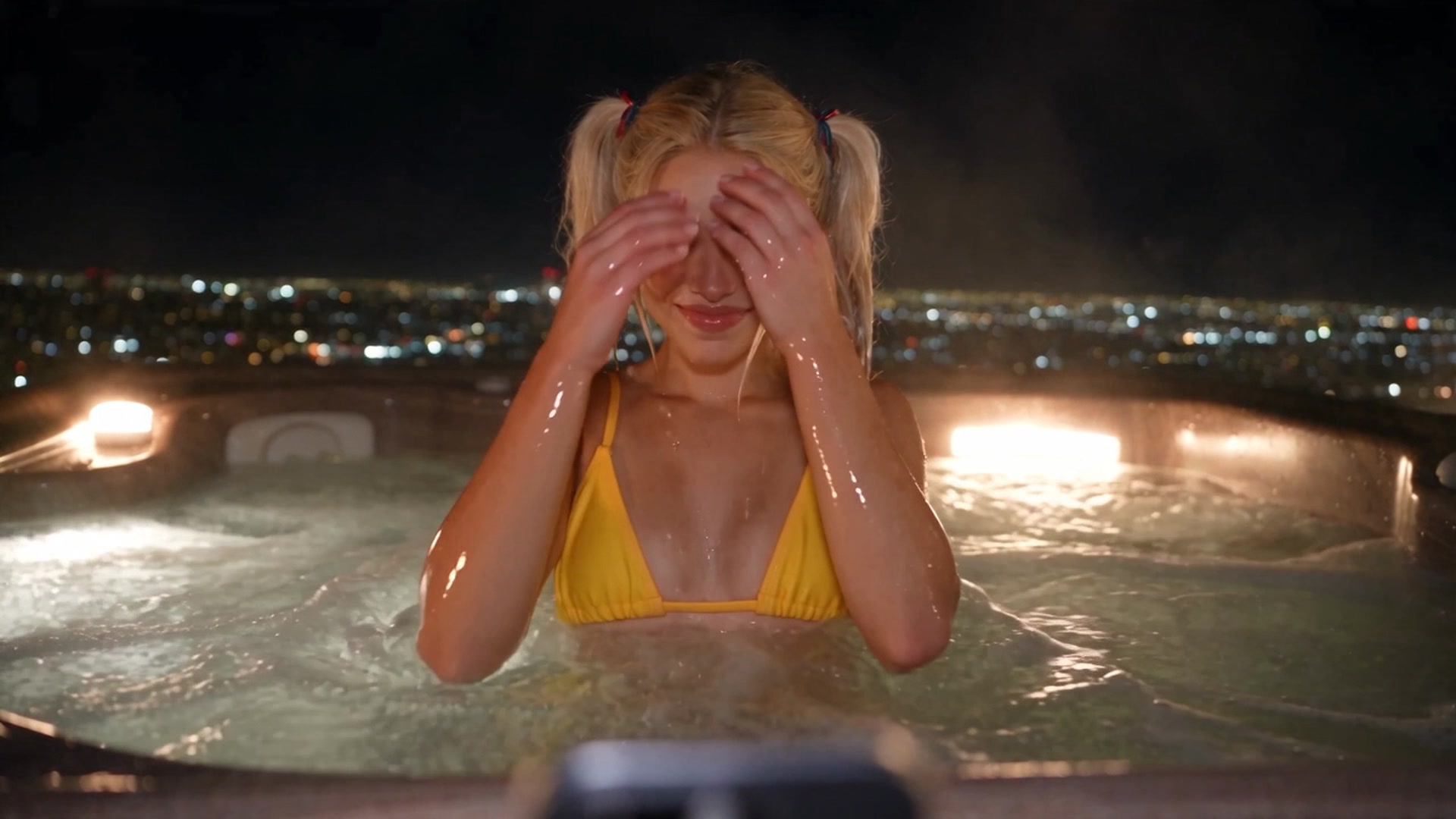 Emely in a yellow triangle bikini top with wet hair in twin ponytails, shielding her eyes with both hands while sitting in an outdoor hot tub at night with a city skyline glowing in the background.
