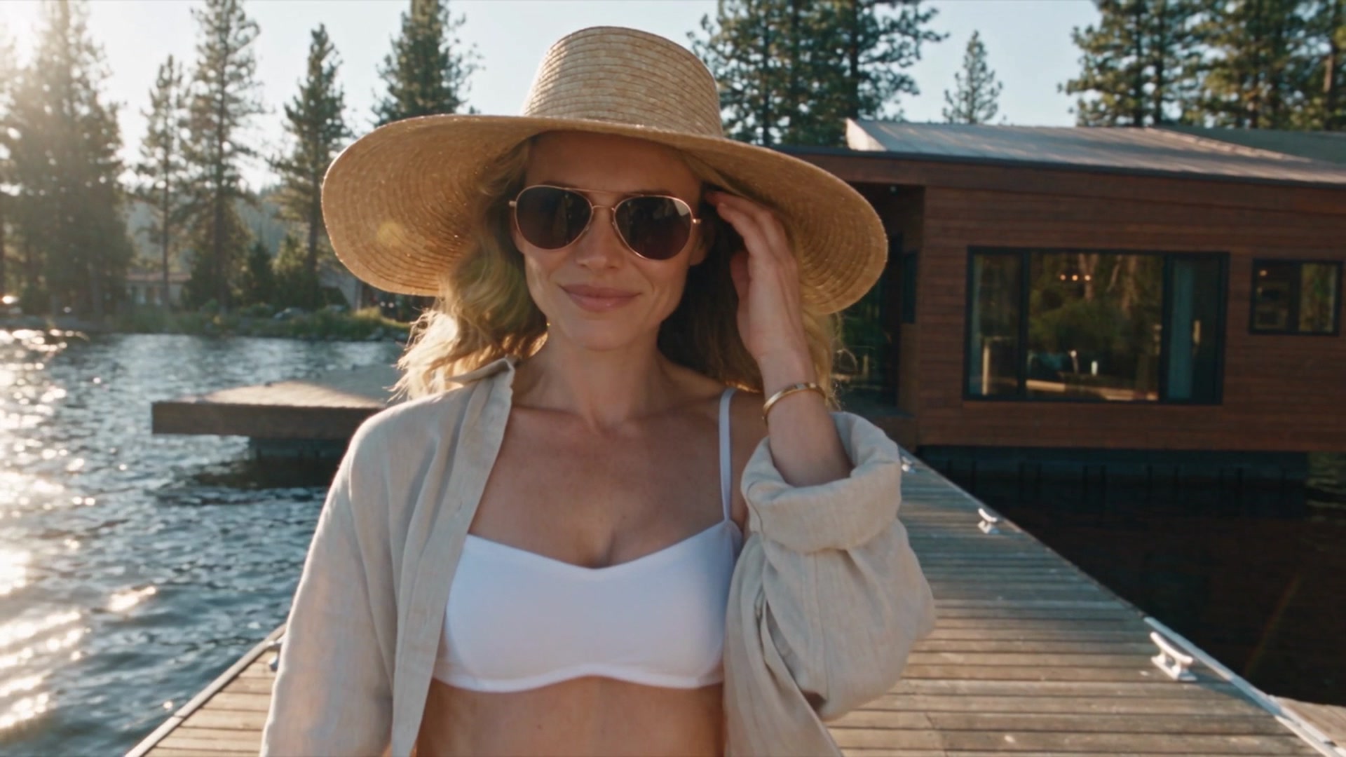 Jade wearing a white bikini top with a beige open shirt and a straw sun hat, standing on a wooden dock by a lake, tipping the brim of her hat while smiling.