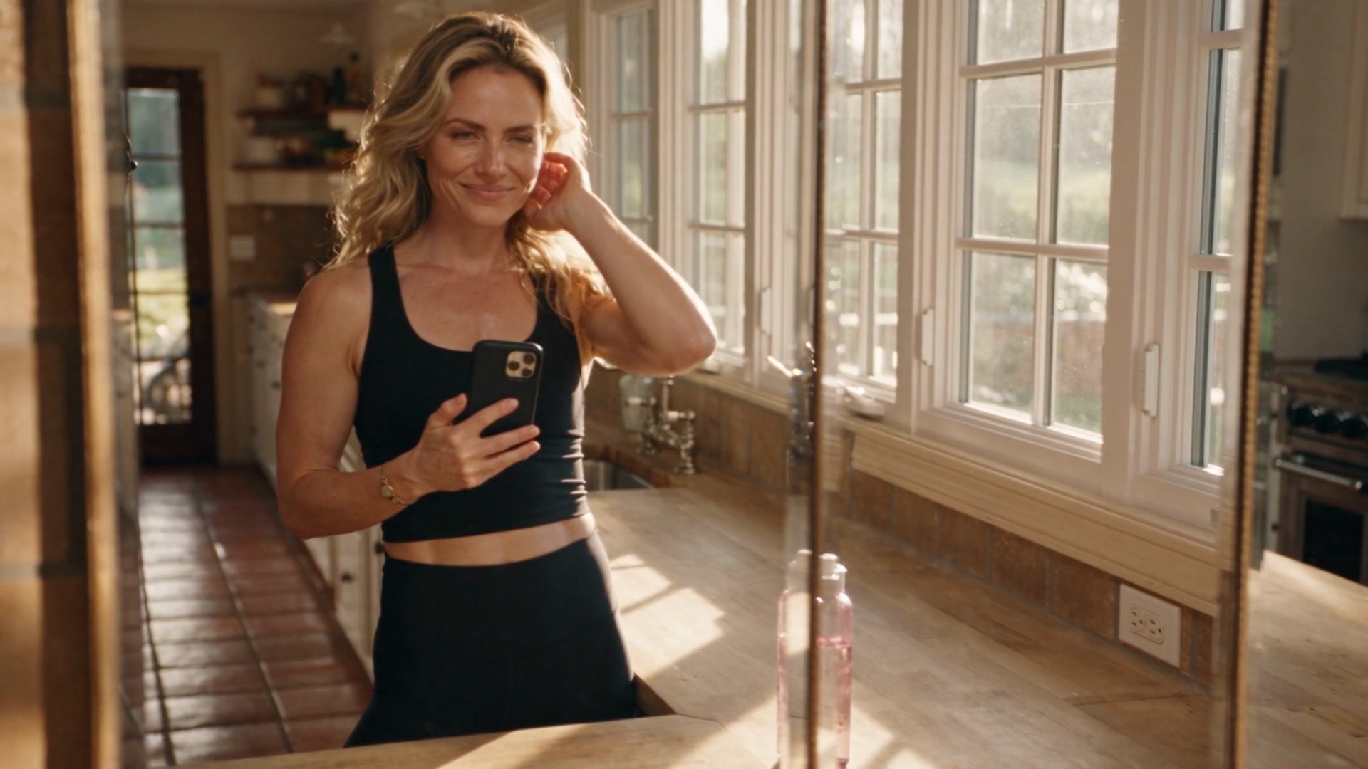 Jade wearing a black athletic tank top and leggings, standing in a sunlit kitchen, taking a mirror selfie with her phone.