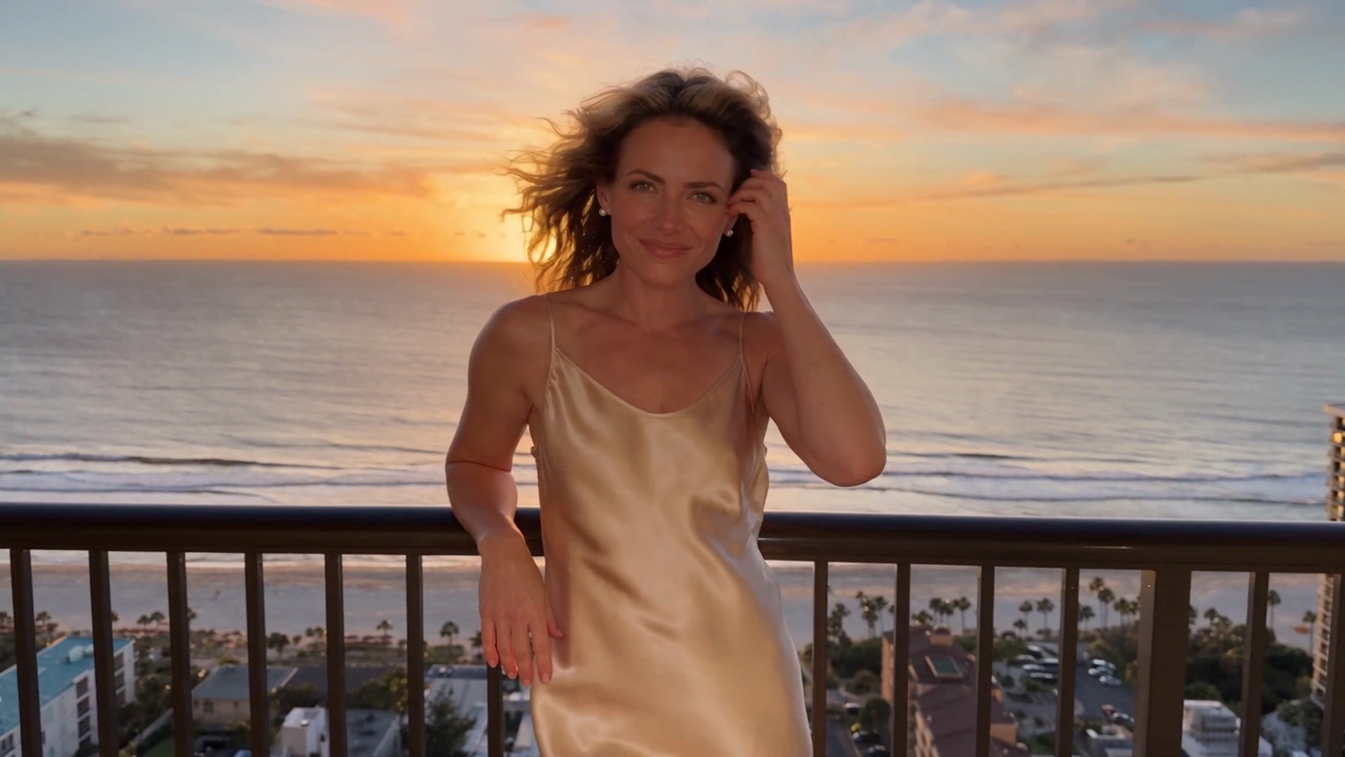 Jade wearing a champagne-colored satin slip dress, leaning against a balcony railing at sunset with the ocean and beachfront skyline behind her.