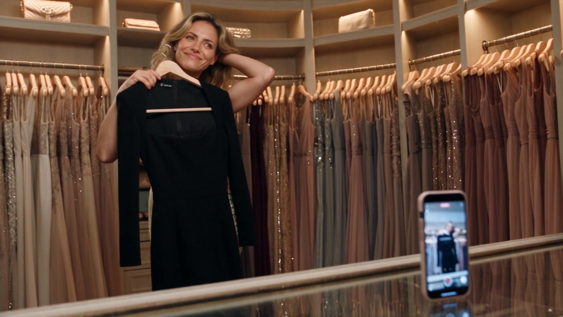 Jade holding a black dress on a hanger up to herself in a walk-in closet lined with rows of gowns, smiling toward a phone propped on the counter.