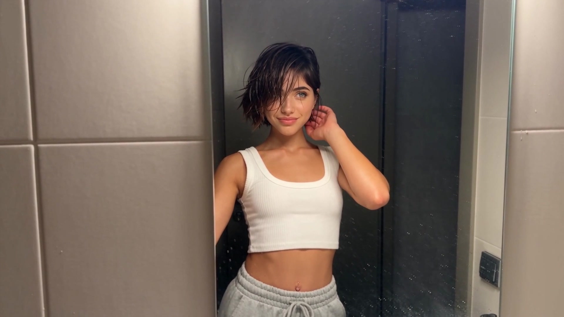 Sofia leaning against a dark glass shower door in a tiled bathroom, hair damp, wearing a white ribbed tank top and grey drawstring sweatpants, one hand tucking hair behind her ear.